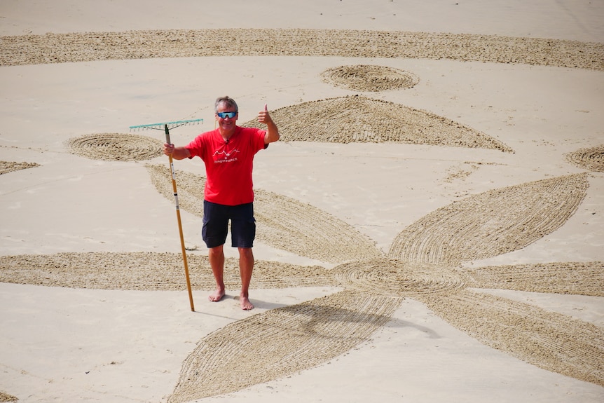 A middle aged man wearing a red t-shirt standing on a beach on a sunny afternoon.