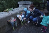 A woman kneels down, holding the hands of two little girls, as they place flowers against a fence. 