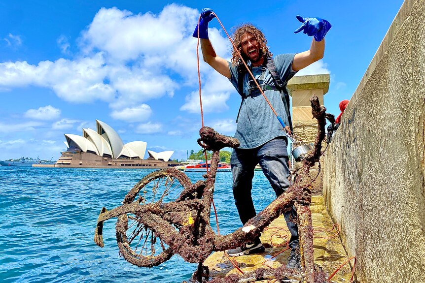 Man with curly hair, uses a magnet to retrieve rusted bike from Sydney Harbour, wears blue gloves, yells, points to the bike.