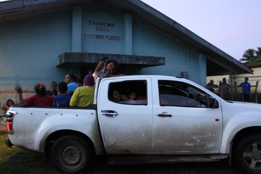 Wide-shot of people smiling and looking-on from the back of, and inside, a ute out Tabwewa Hall in Tabwewa Village, Fiji.