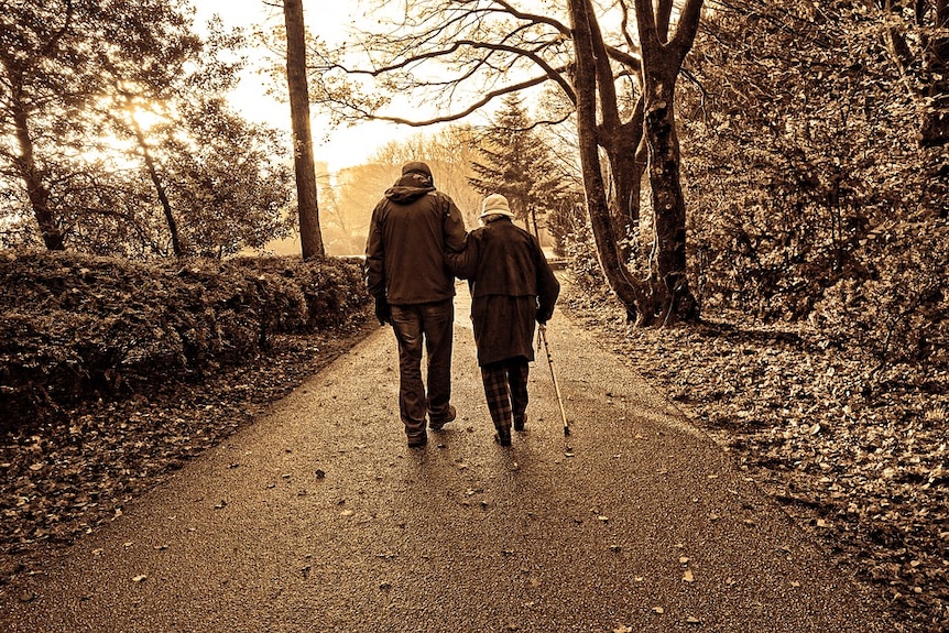 Man and woman walk through a park arm in arm. Woman is holding a walking stick.