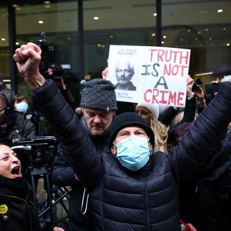 A masked man looks to the camera as he holds both his hands in the air in fists in celebration, in front of other celebrators.