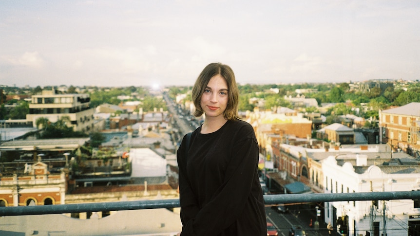 A young woman smiling demurely on a rooftop overlooking town streets