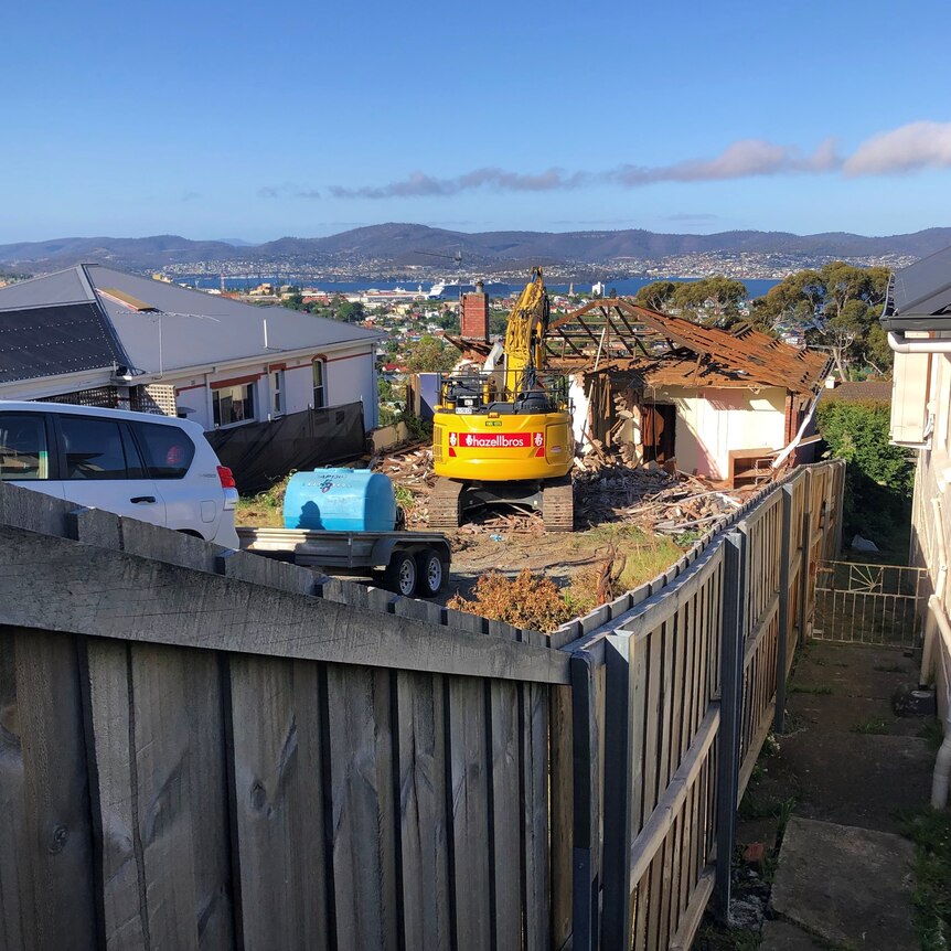 A yellow wrecking crane in front of a partially demolished single-storey home on a hill overlooking Hobart.