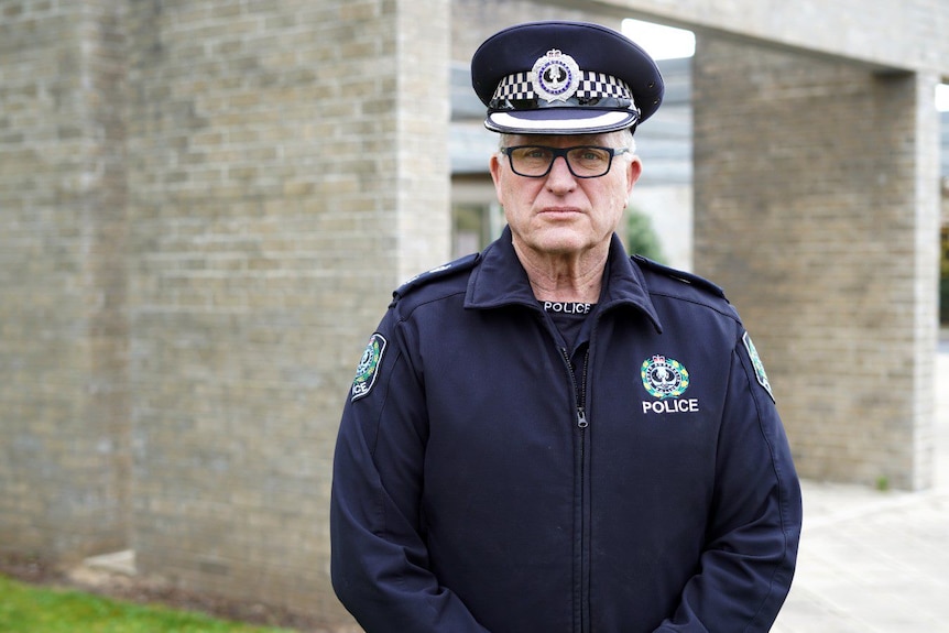 A male police officer in uniform stands in front of a brick structure