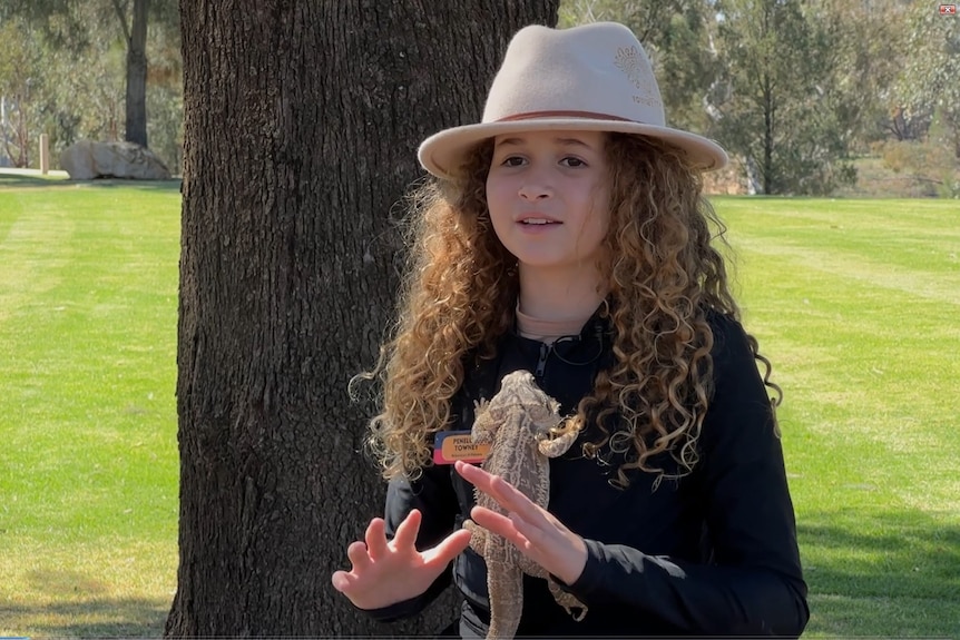 A young girl with long, curly hair, wearing a broad-brimmed hat and cuddling a lizard, talking outside. 