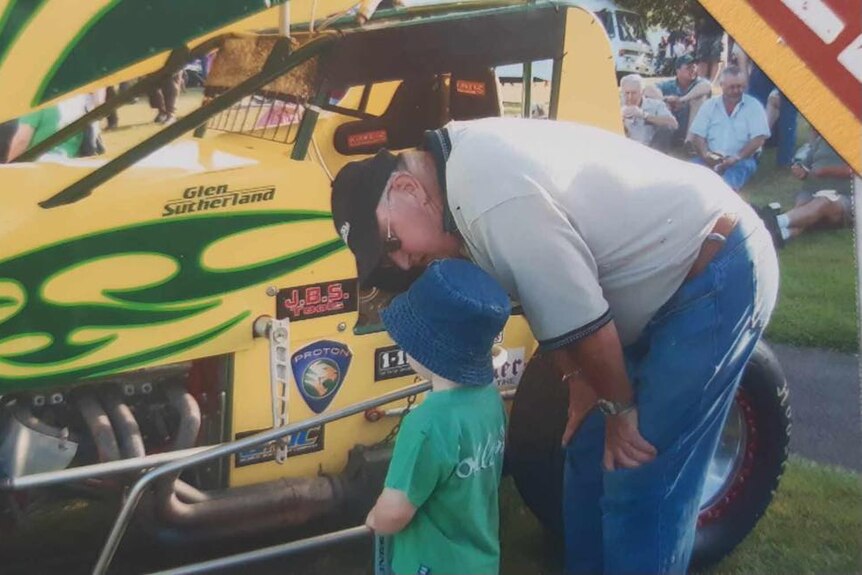 Toddler looks at bright yellow car with green flames painted on as his grandfather leans over to tell him about it.