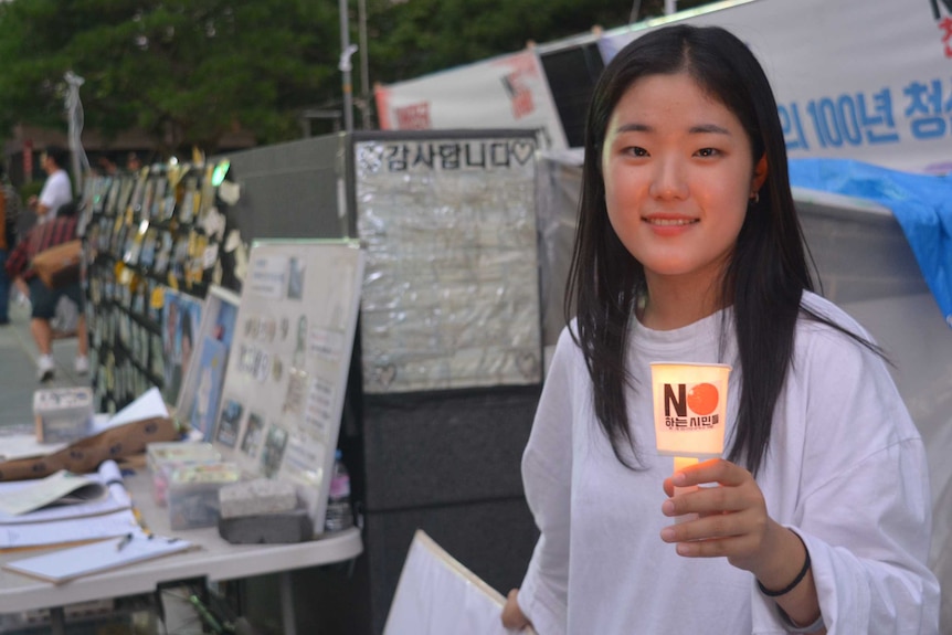 A young woman in a white t-shirt smiles and holds a candle in front of protest signs.