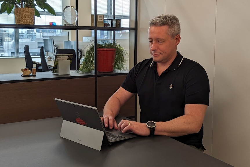 Guy sits at an office table wearing a dark blue polo shirt, typing on a laptop computer.