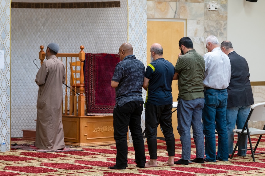 Behind view of Imam leading prayers inside mosque as five men pray behind him.