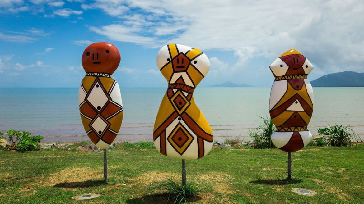 Three large body and head-shaped painted sculptures on metal poles, standing on grass with sea and islands behind.