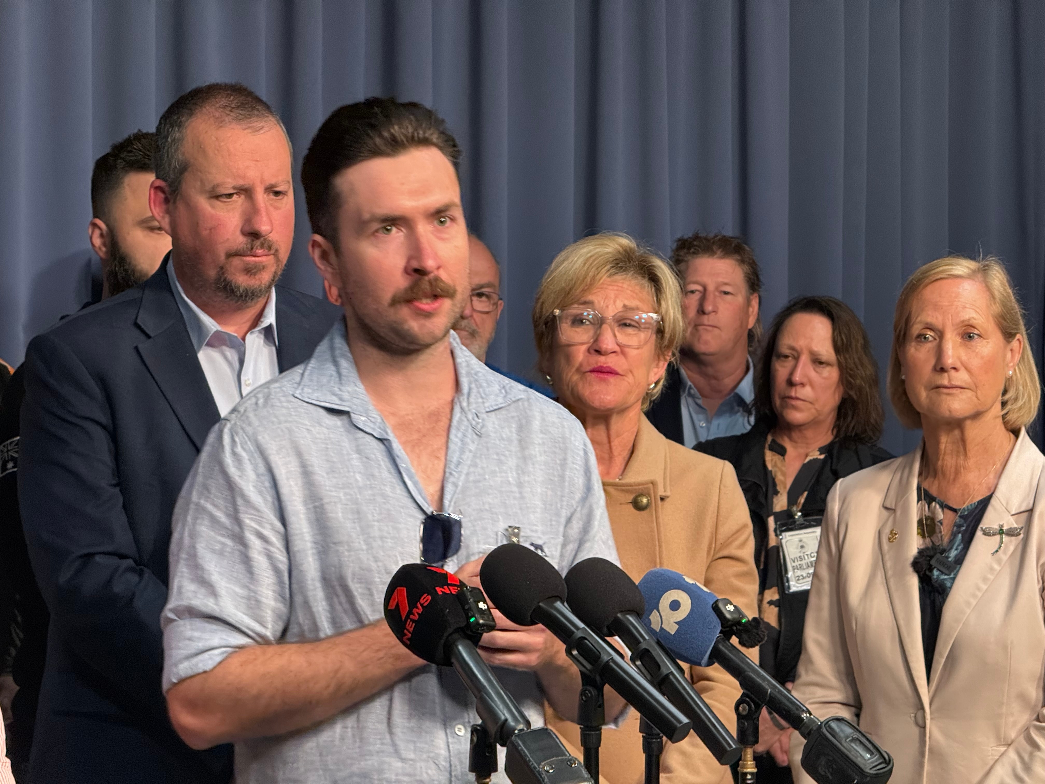 Mr Kirwan stands with his arms clasped in front of media microphones with a serious expression with people behind.