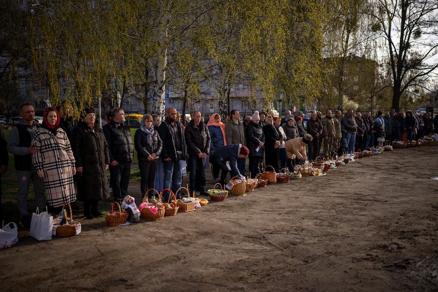 Worshippers stand along a dirt road in a line with baskets of traditional cakes and painted eggs in front of their feet.