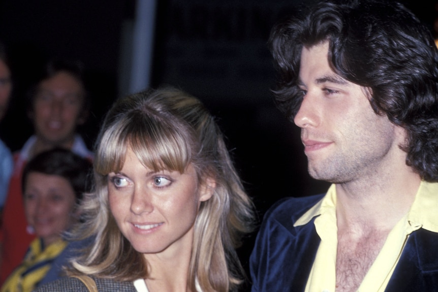 A fair-haired girl with bright eyes and a soft smile standing next to a dark, curly haired man with a three-day growth