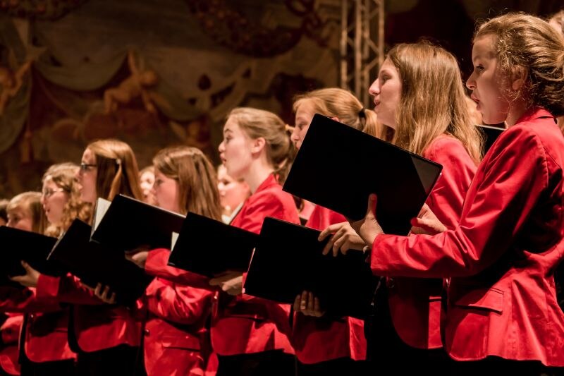 Members of the Hanover Girls Choir performing on stage. Wearing red tops and holding black folders