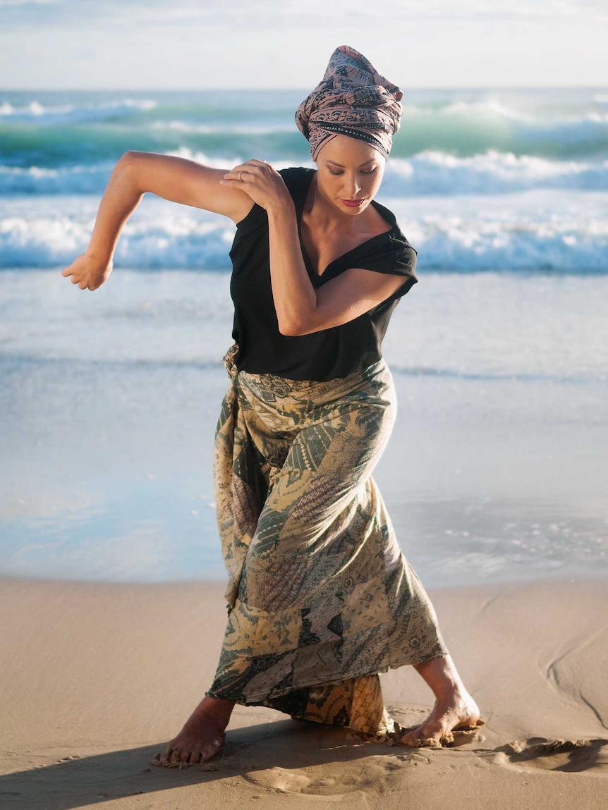 On a bright day, you view a woman in a traditional Indonesian dance pose on the beach.