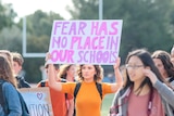 A girl wearing orange stands on a school football field holding a sign which reads: fear has no place in our schools
