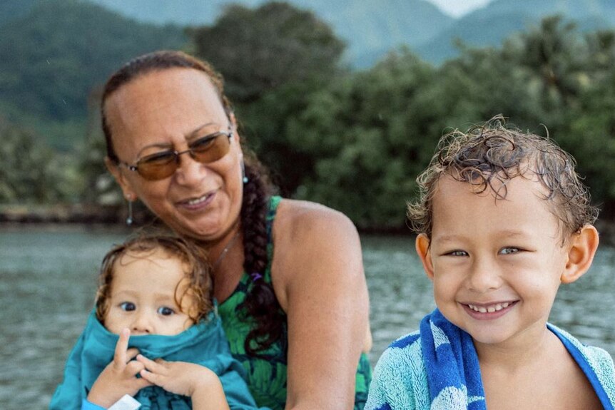 Woman with glasses and green dress sits smiling with her two young grandchildren. Water and trees behind them. 