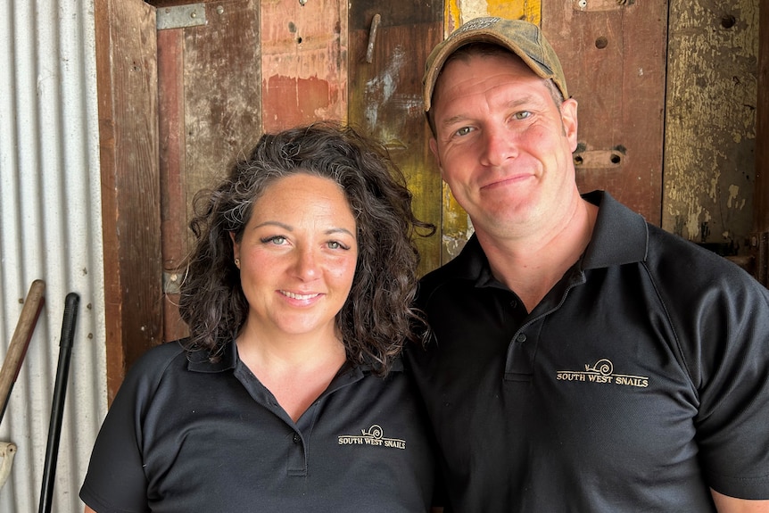 A smiling man and woman stand in front of a wooden barn door both wear black tee with a small logo on left, man wears cap.