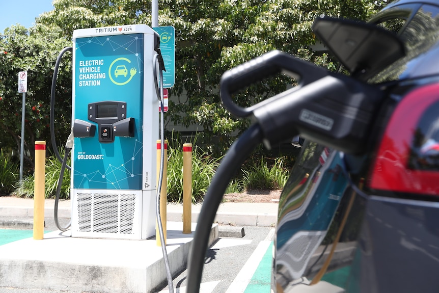 Une console de recharge verte pour véhicules électriques se trouve sur une place de parking en béton, tandis qu'une voiture électrique rouge est chargée au premier plan.