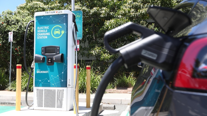 A green electric vehicle charging console sits in a concrete parking spot, as a red electric car is charged in the foreground.