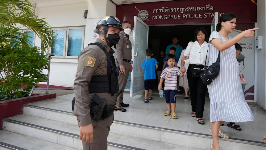 Thai police man stands in khaki uniform, black mask and helmet, as Chinese women and children leave police station.