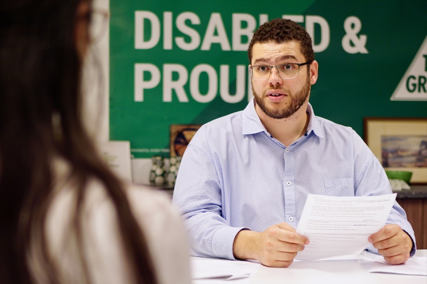 Greens Senator Jordon Steele-John speaks to someone in an office. Behind him, a banner reads "Disabled and proud".