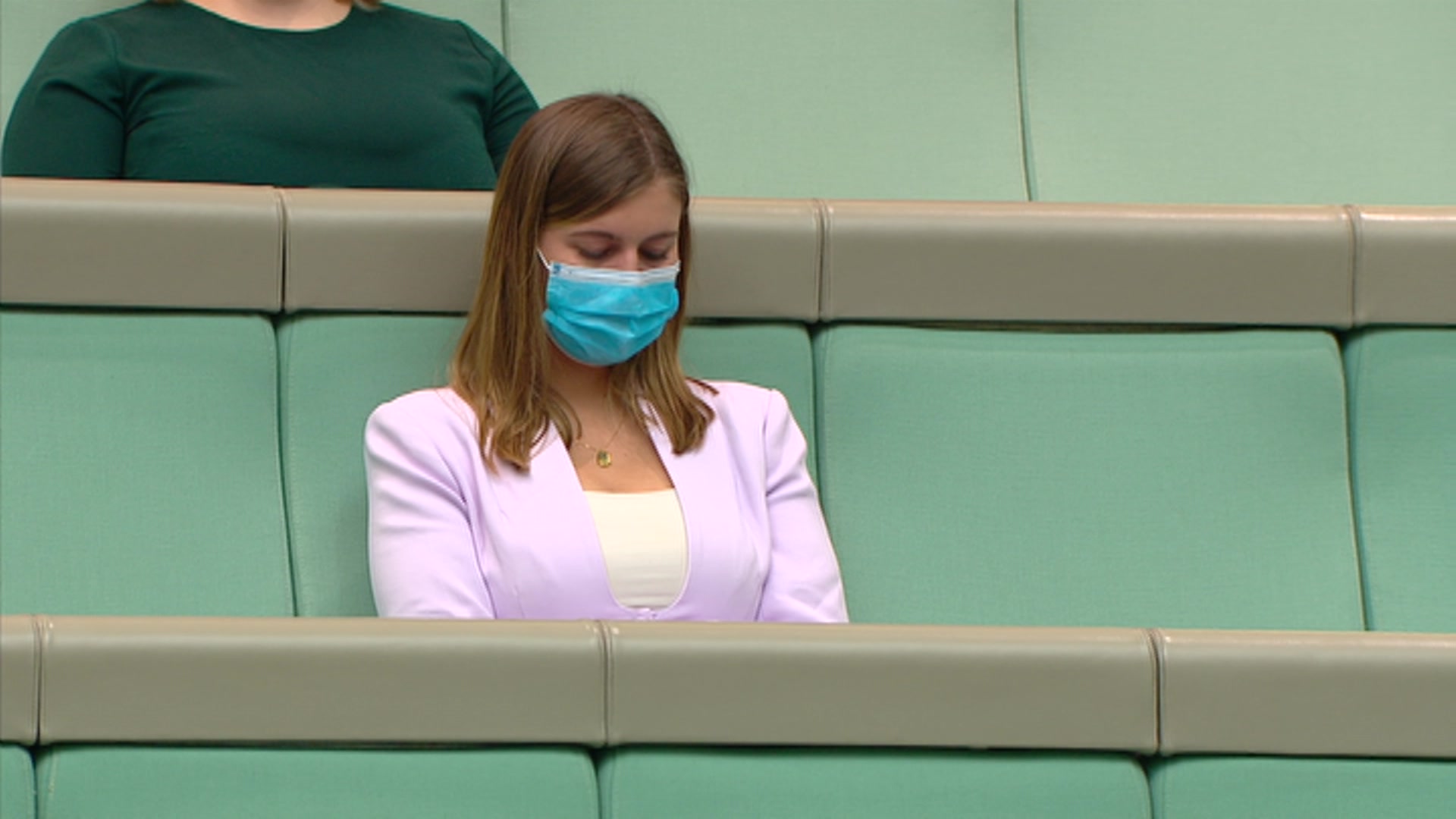 Brittany Higgins wearing a purple blazer and mask sitting in a green seat in the House of Representatives gallery, looking down.