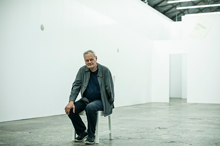 Artist Mike Parr in grey jacket, sits on white chair with right hand on knee inside white gallery space with concrete floor.