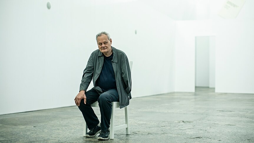 Artist Mike Parr in grey jacket, sits on white chair with right hand on knee inside white gallery space with concrete floor.