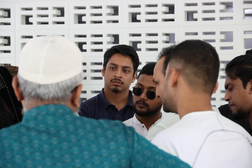 A group of men in traditional Islamic dress standing and speaking, outside in front of a wall.