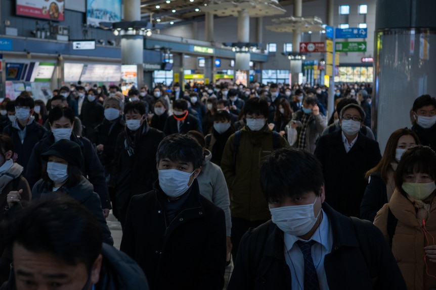 A crowd of Japanese people, all wearing face masks, at a railway station.