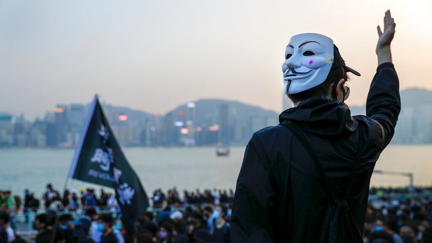 You view the back of a black-clad protester with a Guy Fawkes mask as he raises his right arm in front of a crowd.