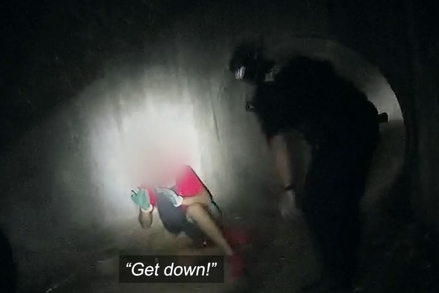Police shine a flashlight at a young person who is sitting on the ground with their hands raised