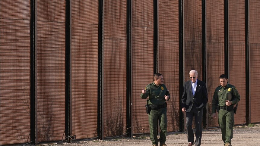 Mr Bident walks beside a fence that stretches across the whole photo next two men in uniform. 