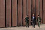 Mr Bident walks beside a fence that stretches across the whole photo next two men in uniform. 