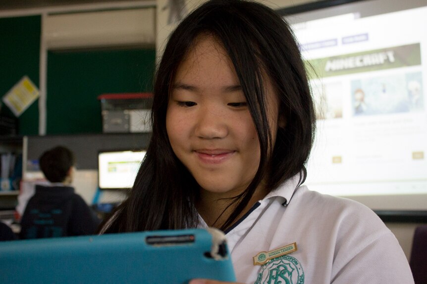 A girl uses a tablet, large computer display in background.