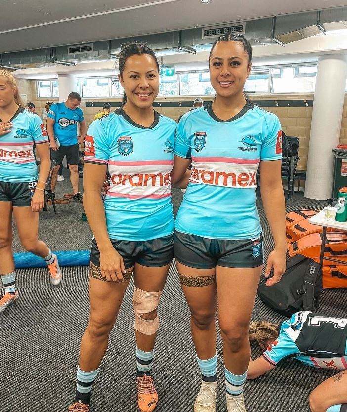 Two sisters dressed in blue rugby uniform smile to camera in locker room. 