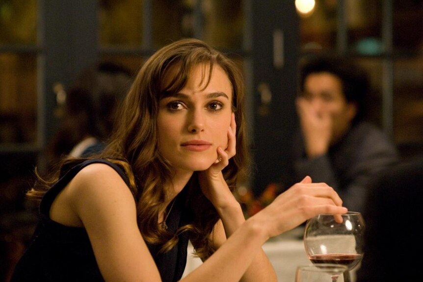 Actor Keira Knightley in a movie scene where she is seated at a fine dining table with her hand on a glass of red wine