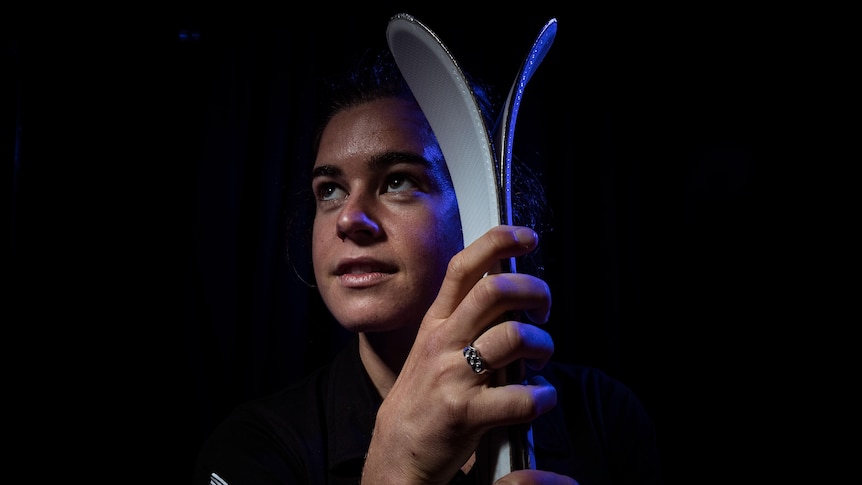 Britt Cox poses with her skis in front of a black background and looks to the side.