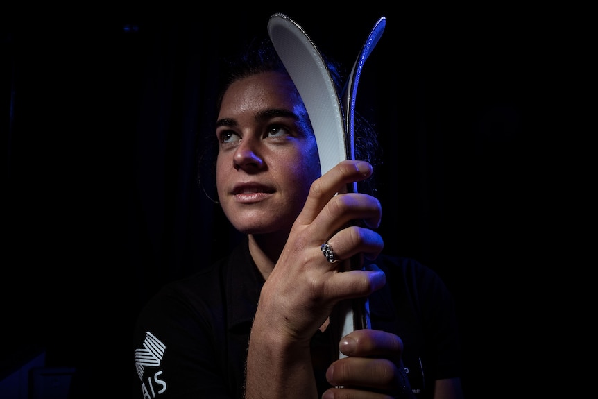 Britt Cox poses with her skis in front of a black background and looks to the side.