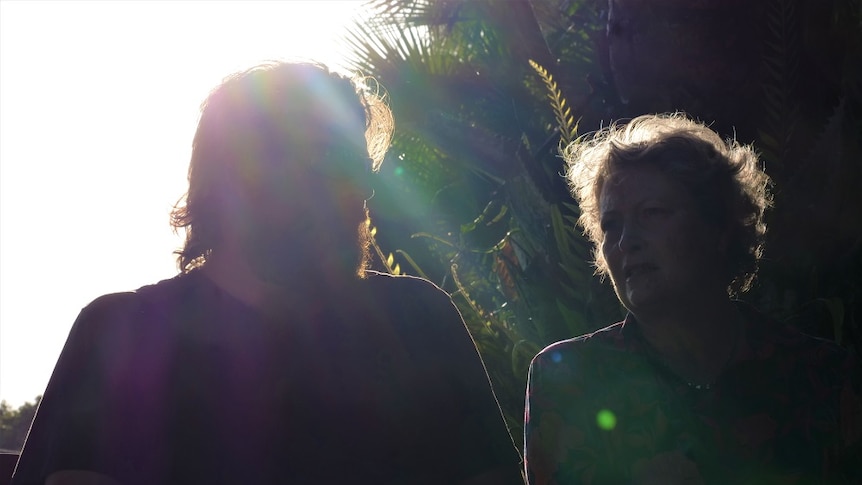 Silhouette image of mother and adult son sitting on park bench with sun and palm trees in background.