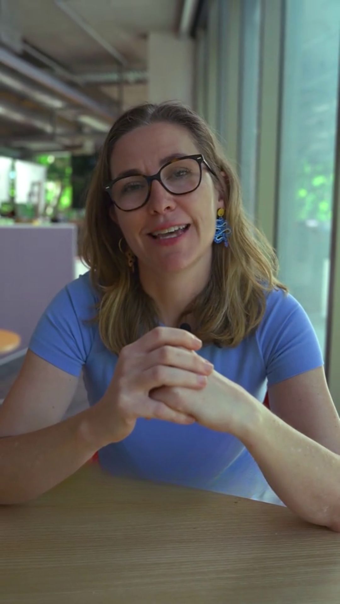 A blonde woman in a blue shirt and glasses sits at a table with her hands clasped. 