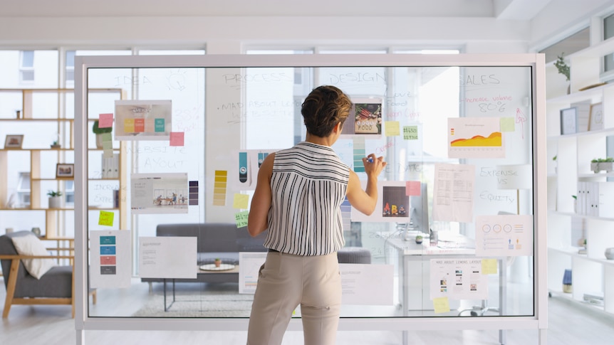 woman in modern office brainstorms on glass wall with marker and post it notes