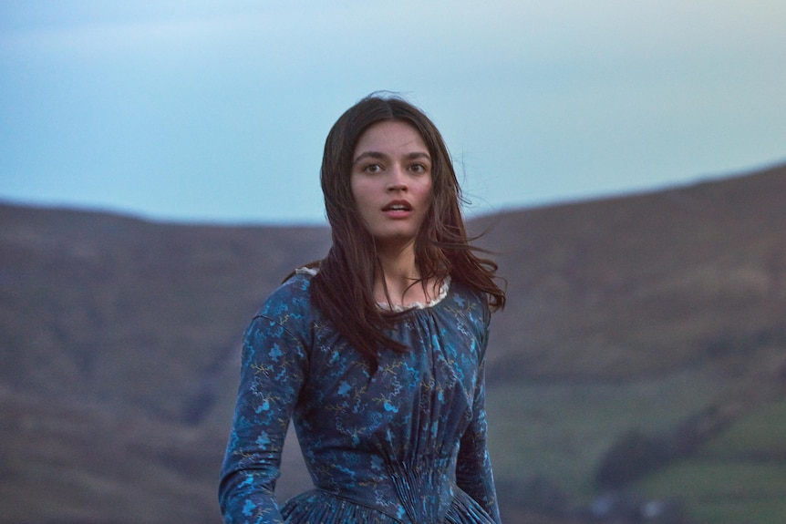 A young white woman with brown hair wearing a blue regency style 1800s dress walks through windy moors.