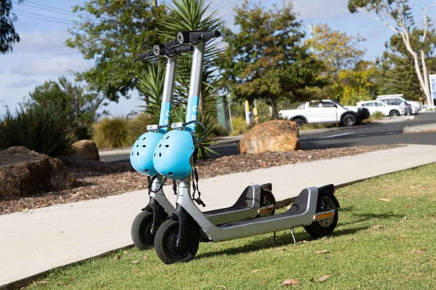 E-scooters with blue helmets on the grass and footpath behind and car park further behind