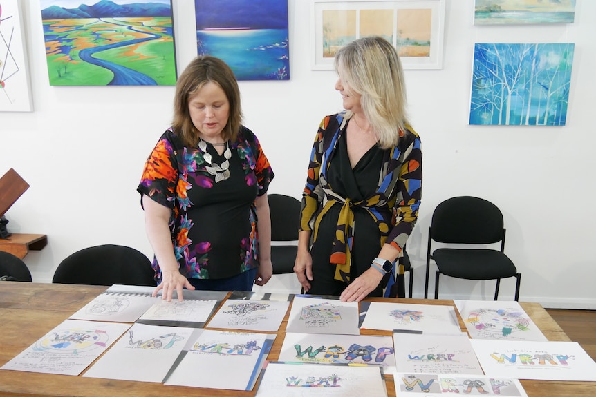 Two women in flowered shirts stand in an art gallery and look at works of art on a table. 