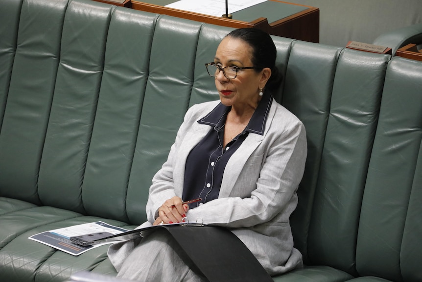 Linda Burney reacts in Parliament. She is an Indigenous woman with black hair tied in a bun, wearing a grey suit and glasses. 