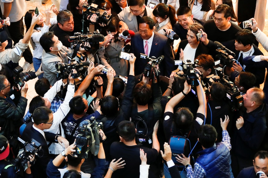 A crowd of people holding cameras and microphones surrounds a man dressed in a suit.