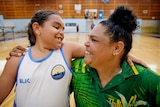 A young girl and her mum smile happily at each other. They are in a basketball court.